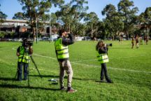 Three students on the side of an AFL field, wearing fluorescent vests taking film and photo content of the game playing. 